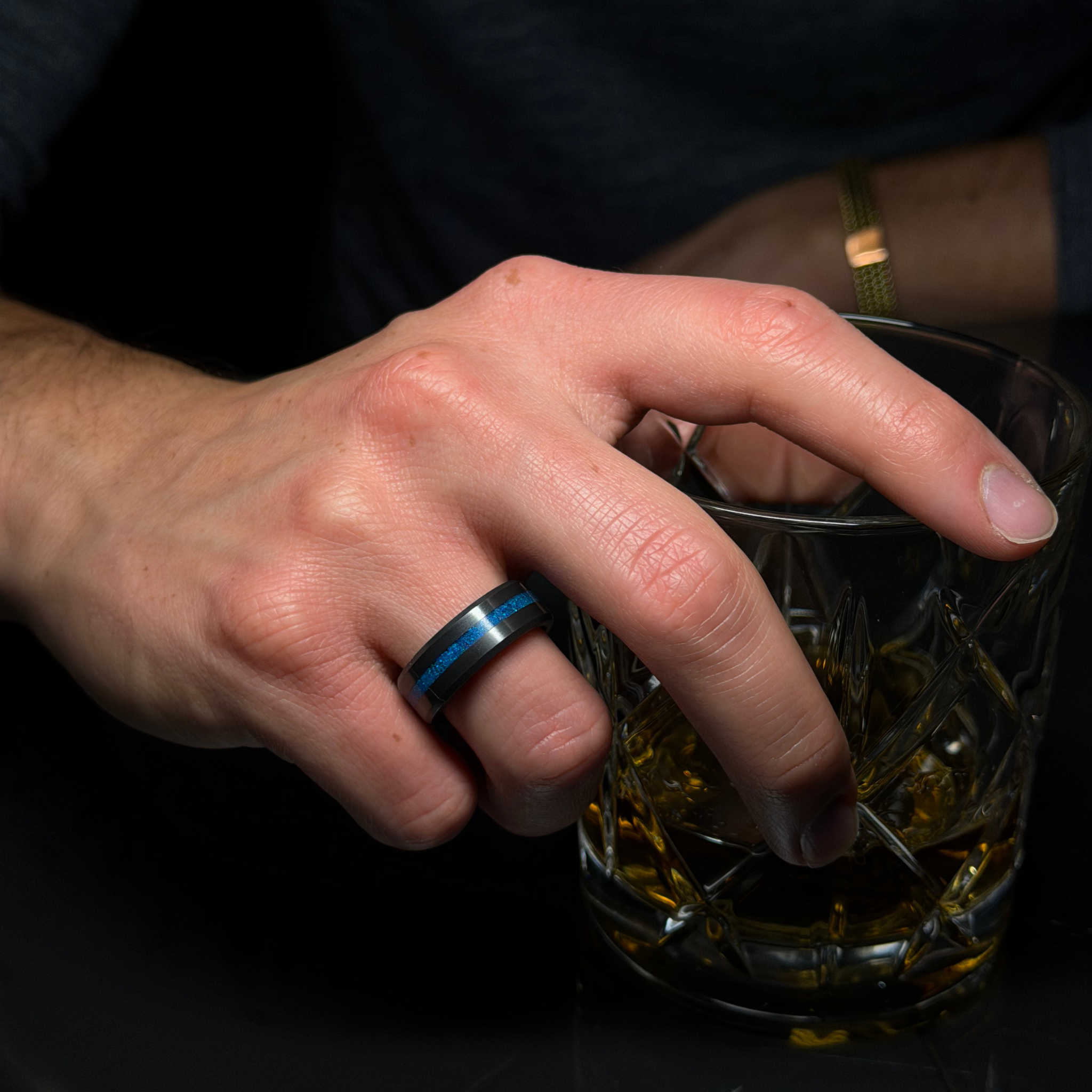 Hand wearing a solid black diamond ring with a blue opal inlay, holding a glass of whiskey on a dark background